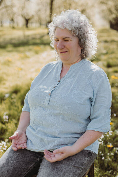 Carola Harter Channelmedium für Engel und die Geistige Welt, Engelmedium in Oberkirch sitzt in einer grünen Obstbaumwiese mit einer blauen Bluse in und lächelt mit geschlossenen Augen in eine Meditation versunken
